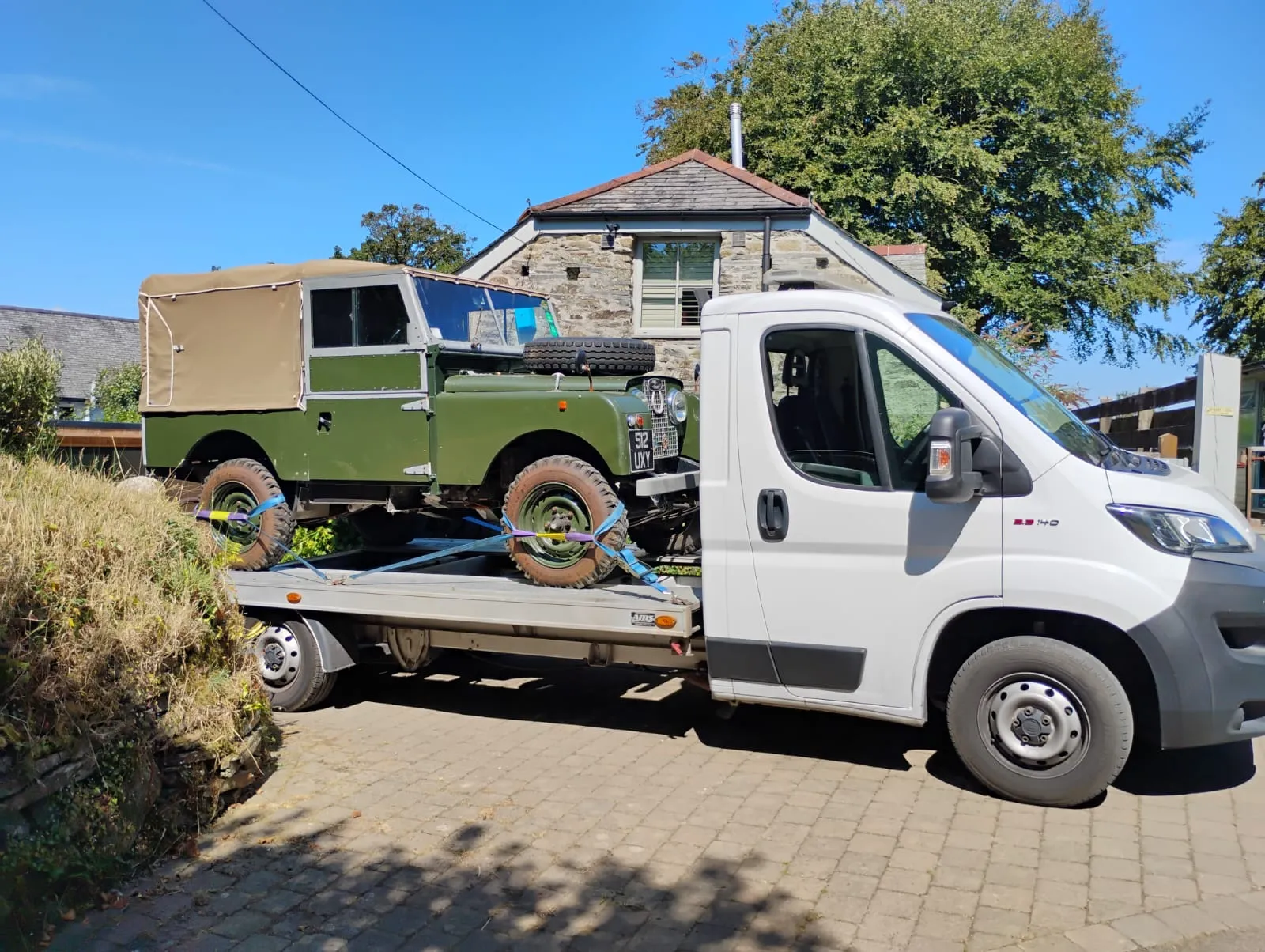 Classic green Land Rover Series 1 strapped onto the transporter outside a stone cottage